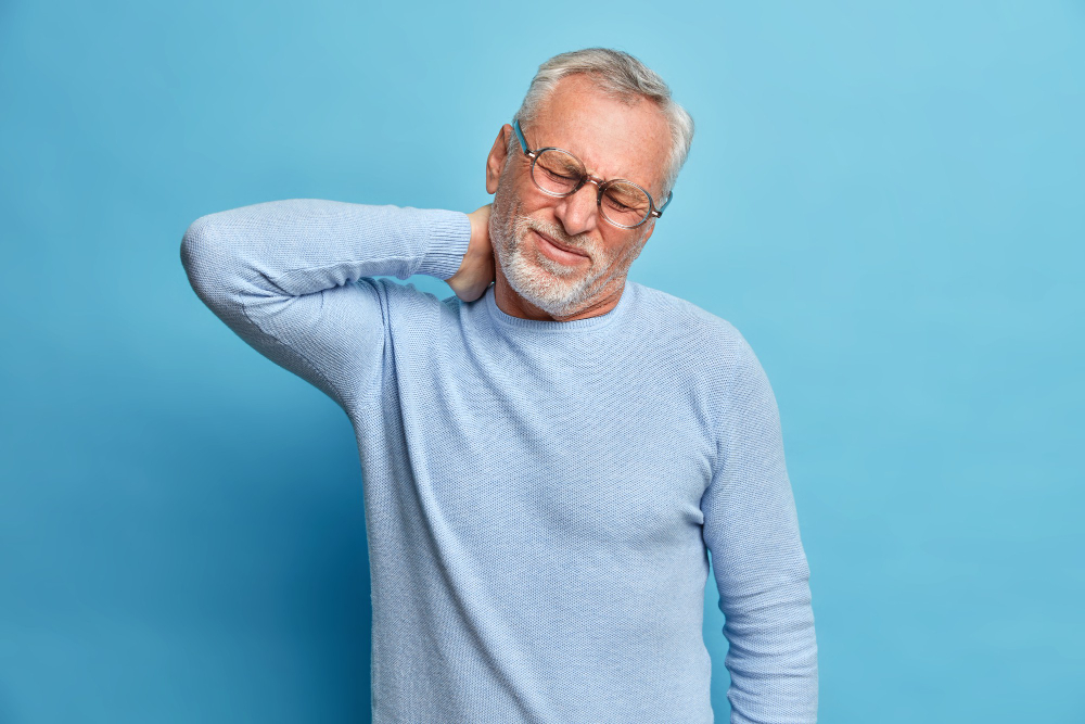 Free photo aged exhausted bearded european man touches neck suffers from pain in neck tilts head grimaces from painful feelings needs massage dressed in long sleeved jumper isolated over blue wall
