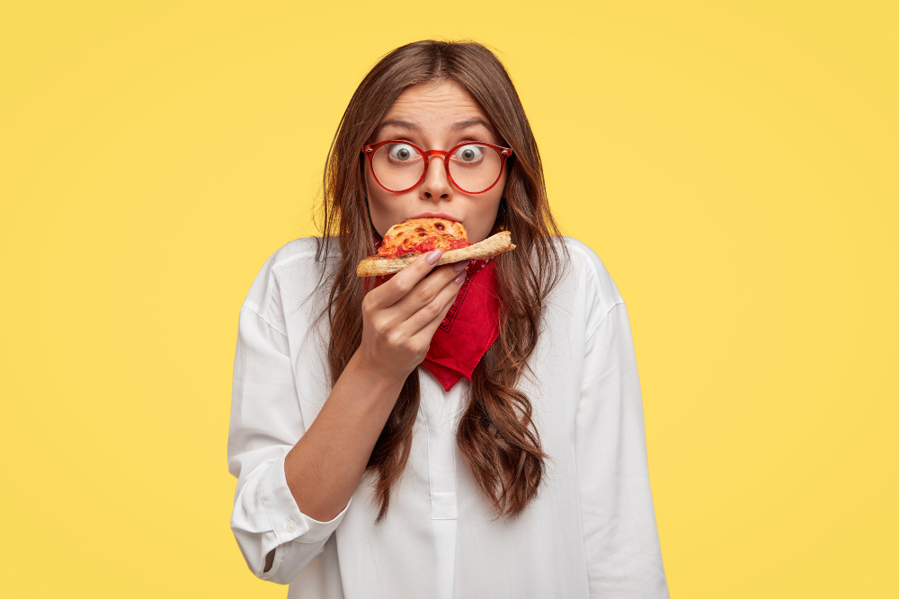 Free photo surprised european fashionable woman has slice of pizza, looks dressed in oversized shirt, surprised with very nice taste, isolated over yellow wall. people and fast food concept
