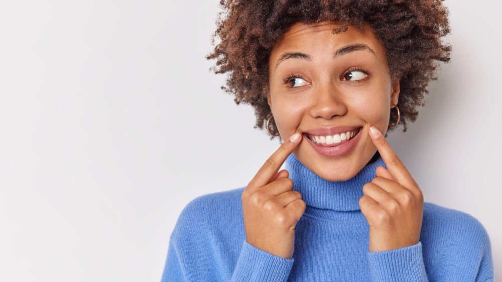 Free photo woman smiles broadly points at her perfect even teeth indicates at corners of lips looks away wears casual blue jumper isolated on white with blank copy space.

