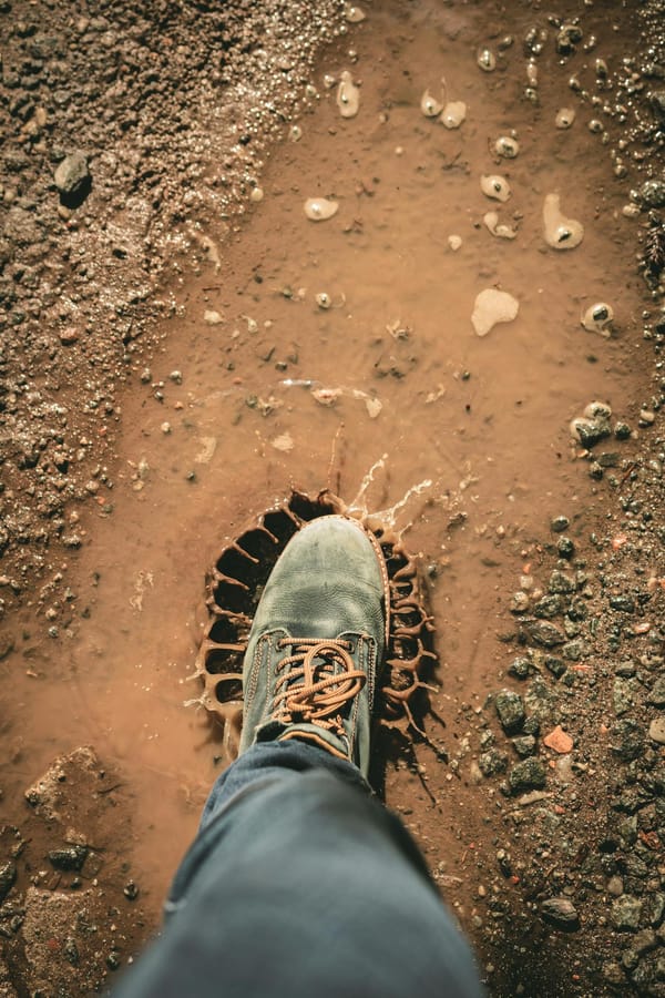 A single boot stepping into a muddy puddle outdoors, creating a splash on a dirt road.