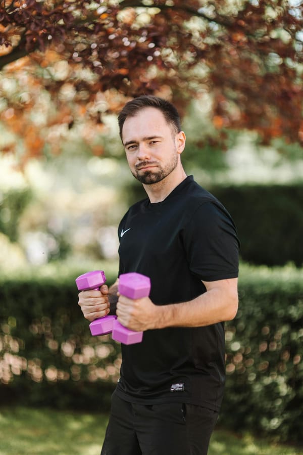 Man in black activewear lifting purple dumbbells outdoors in a garden setting.