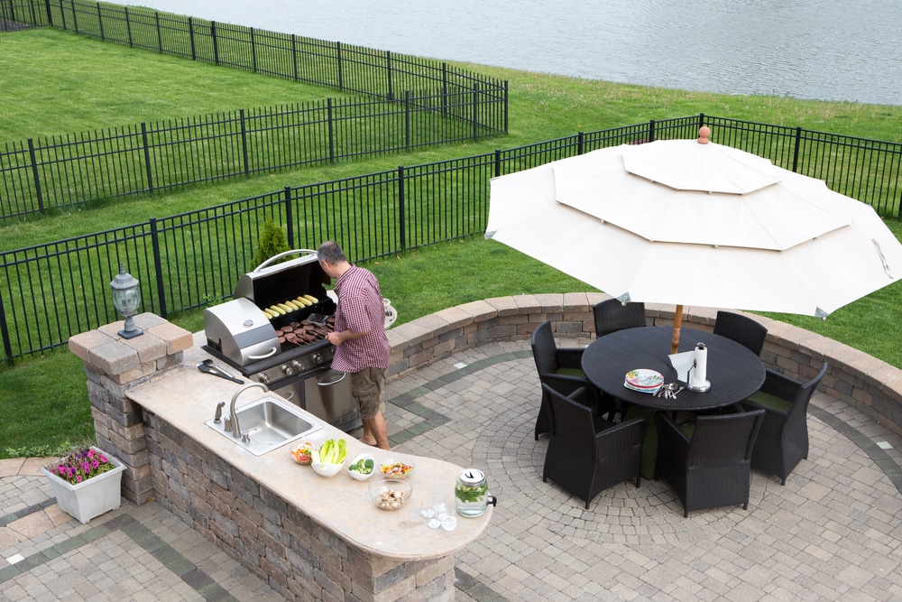 High angle view of a man cooking meat on a gas BBQ standing in the sunshine on a paved outdoor patio at the summer kitchen preparing for guests with a table and chairs with a garden umbrella alongside