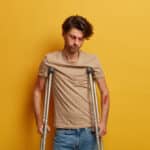 Sad unhappy man looks down, has serious injury after falling down from height, tired of long recovery period, tries to walk with crutches, poses against yellow wall. handicapped disabled male