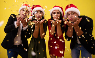 Four friends in christmas hats are partying on the new year's eve, laughing, smiling and throwing vivid golden confetti in the air.