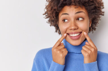 Free photo woman smiles broadly points at her perfect even teeth indicates at corners of lips looks away wears casual blue jumper isolated on white with blank copy space.