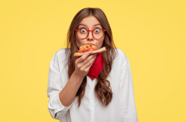 Free photo surprised european fashionable woman has slice of pizza, looks dressed in oversized shirt, surprised with very nice taste, isolated over yellow wall. people and fast food concept