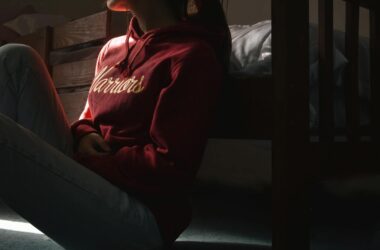 Contemplative young woman in hoodie sitting on the floor in dimly lit room, symbolizing solitude and introspection.
