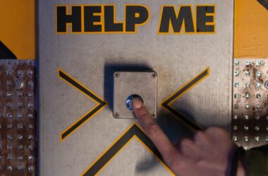 A hand presses an emergency 'help me' button on a metallic panel, evoking sci-fi themes.
