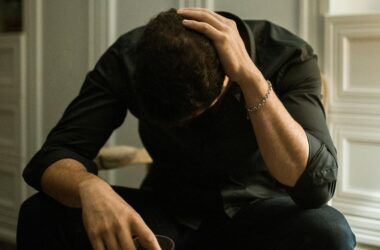 Man in distress seated indoors, head in hand, holding a drink, reflecting solitude.