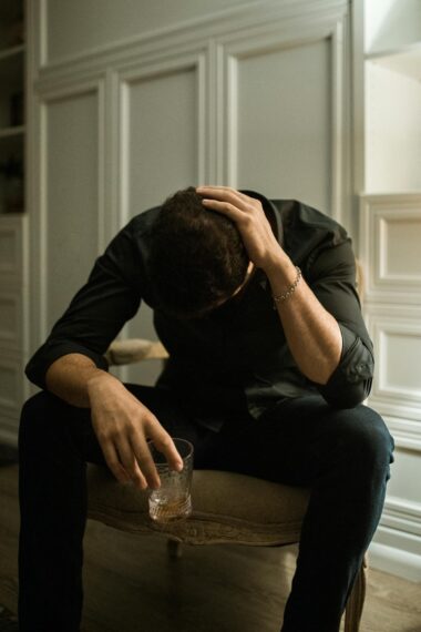 Man in distress seated indoors, head in hand, holding a drink, reflecting solitude.